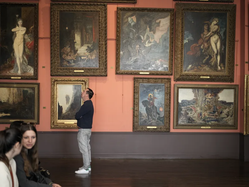 A man standing alone in a museum gallery, looking upward at a large painting while other visitors sit in the foreground 