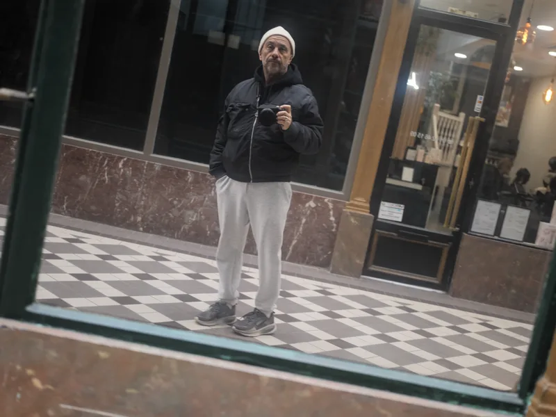 A middle-aged man, camera in hand, photographing himself in a shop window reflection within what appears to be a Parisia