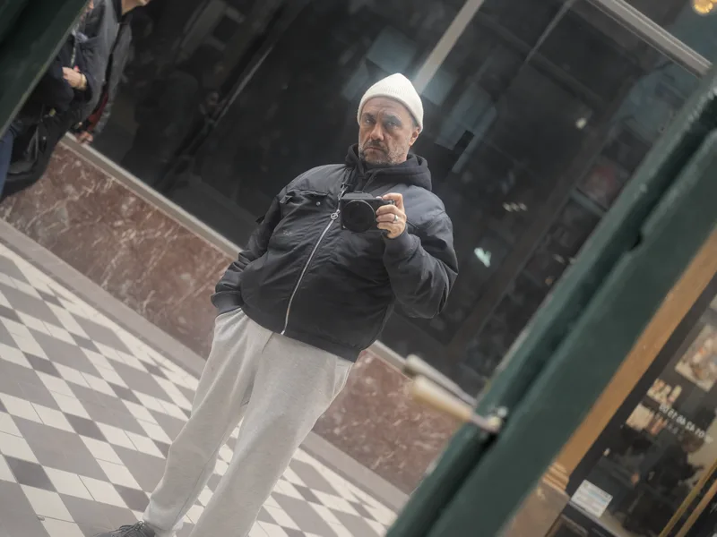 A middle-aged man in a white knit beanie, dark puffer jacket, and light grey sweatpants, holding a compact camera at che