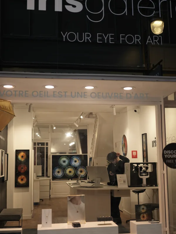 A gallery worker or attendant alone inside an iris photography gallery at night, viewed from the street through the shop