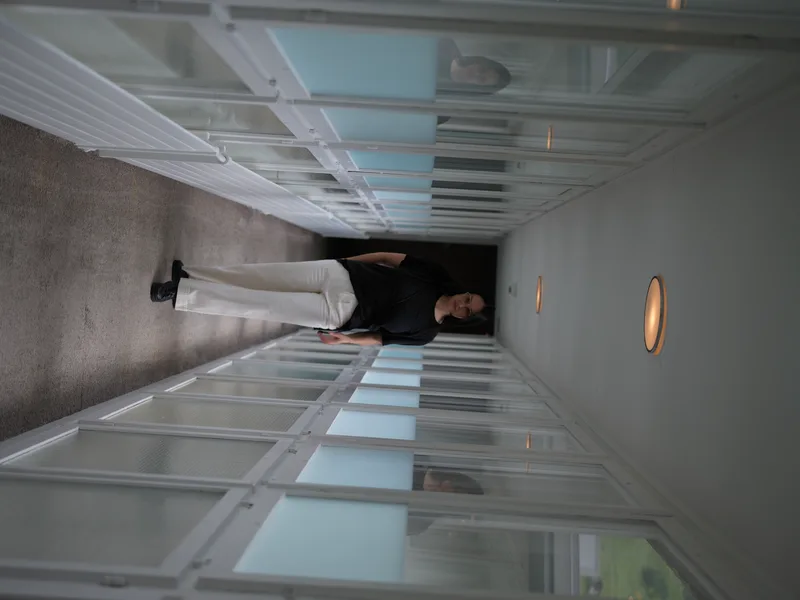 A woman in a black top and white wide-leg trousers, walking toward the camera through a long institutional corridor line