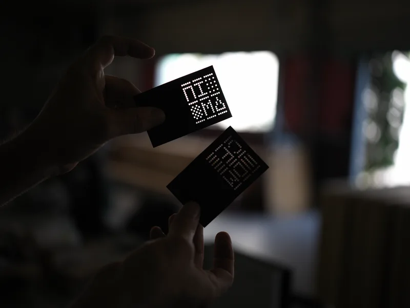 Two black business cards with dot-matrix perforated or illuminated text, held by two hands in low ambient light