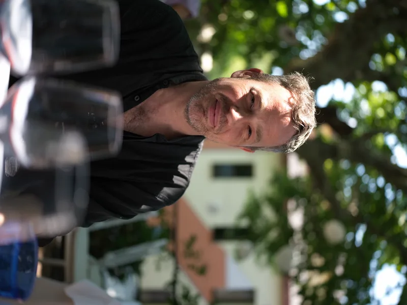 A middle-aged European man, casually dressed in black, seated at an outdoor table with wine glasses in the foreground.